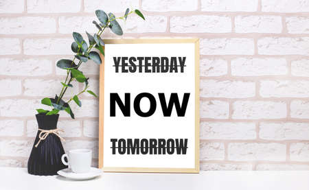 On a white table against a light brick wall, a branch of eucalyptus in a dark vase, a white cup and a light wooden frame with the text YESTERDAY NOW TOMORROW. Home office interior.の写真素材