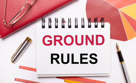 On the desktop is a white notebook with the text GROUND RULES, a pen, burgundy and red tables, and gold-framed glasses. Business concept.の写真素材