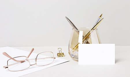 On the desktop are gold glasses, paper clips, pens in a glass beaker, and a white blank card with space to insert text. Business template. Home Office. Close-up of the workplace.の写真素材