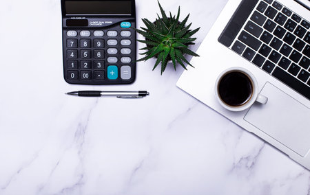 There is a laptop, glasses, a pen, a calculator, a cup of coffee and a green plant on the working...