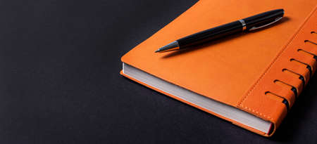 On a black background, an orange notepad and a black pen. copy spaceの写真素材