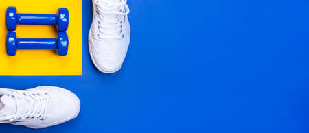 A pair of white sneakers and blue dumbbells on a blue and yellow background. The concept of a healthy lifestyle. view from above. copy spaceの写真素材