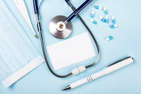 On a light blue background, a stethoscope, a medical mask, pills, a pen and a white card with a...