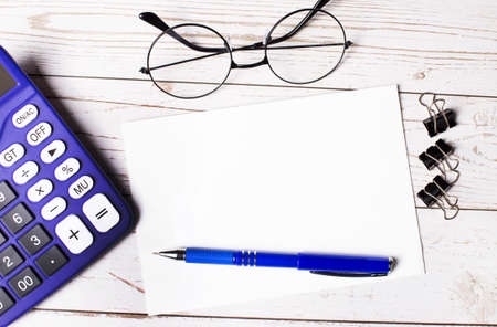 On the wooden desktop is a blue calculator, paper clips, glasses, a blue pen and an empty notepad with space to insert text. copy spaceの写真素材