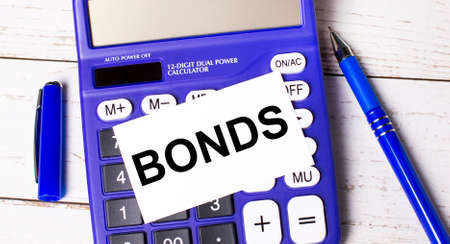 A large blue financial calculator, a blue pen and a blank card with the text BONDS on a light wooden background. Concept of calculations, accounting, calculations, profit.の写真素材