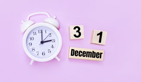 Two wooden cubes and a block with the text DECEMBER 31 and a white alarm clock on a light pink background.の写真素材