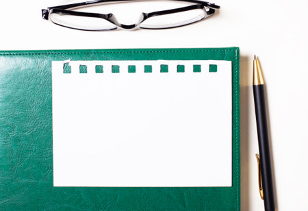 On a light background, glasses, a pen, a green notepad and a white blank sheet of paper with a place to insert textの写真素材