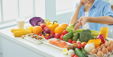 happy young housewife sitting in the kitchen preparing food from a pile of diverse fresh organic fruits and vegetables, selective focusの写真素材