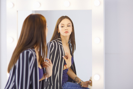 Amazing young woman doing her makeup in front of mirror. Portrait of beautiful girl near cosmetic tableの写真素材