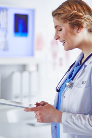 Young woman doctor is standing with board with clipboard smiling in hospital officeの写真素材