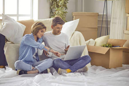 Portrait of happy couple looking at laptop computer together sitting in new house, surrounded with boxesの写真素材