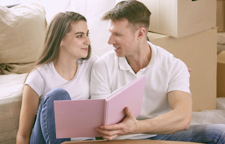 Portrait of happy couple looking at laptop computer together sitting in new house, surrounded with...