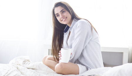 Young beautiful happy brunette woman in black silk dressing gown lying in bed, drinking morning coffee, white cup. Womens day holidayの写真素材