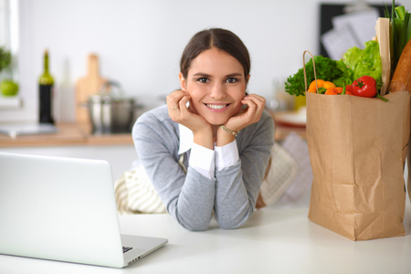 Beautiful young woman cooking looking at laptop screen with receipt in the kitchenの写真素材