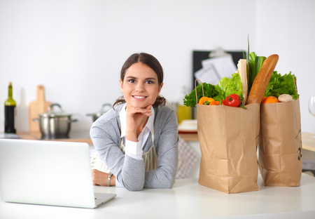 Beautiful young woman cooking looking at laptop screen with receipt in the kitchenの写真素材