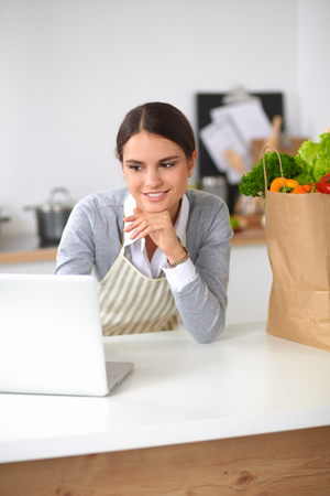 Beautiful young woman cooking looking at laptop screen with receipt in the kitchenの写真素材