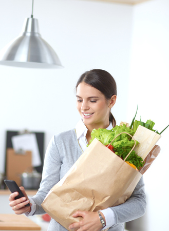 Smiling woman with mobile phone holding shopping bag in kitchenの写真素材