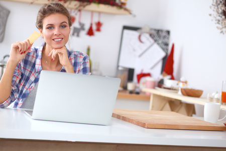 Smiling woman online shopping using computer and credit card in kitchenの写真素材