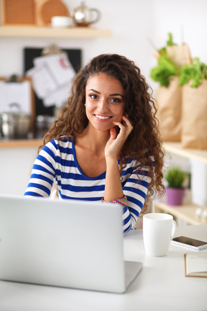 Smiling young woman with coffee cup and laptop in the kitchen at home .の写真素材