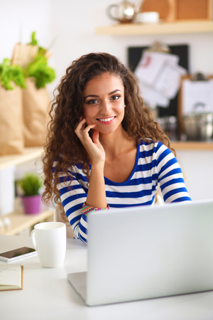 Smiling young woman with coffee cup and laptop in the kitchen at home .の写真素材