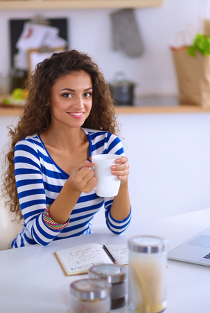 Smiling young woman with coffee cup and laptop in the kitchen at home .の写真素材