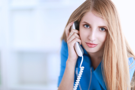 Young woman doctor in white coat at computer using phone.の写真素材