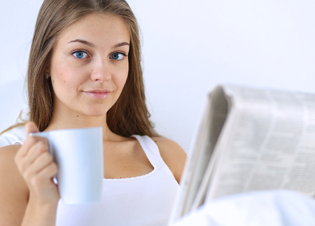 A pretty young woman reading the newspaper in bed and enjoying a cup of tea.の写真素材