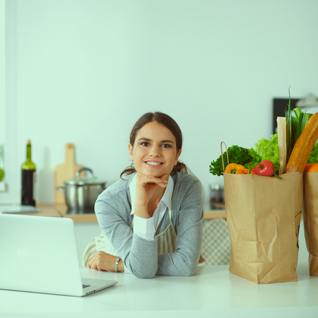 Beautiful young woman cooking looking at laptop screen with receipt in the kitchenの写真素材
