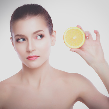 Beautiful young shirtless woman holding piece of orange in front of her eye while standing against white backgroundの写真素材