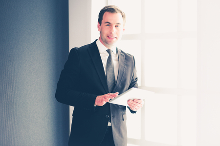 Portrait of young businessman in office with big window. Businessman using tablet computer and looking at cameraの写真素材