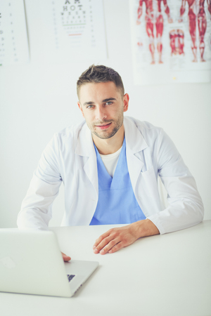 Portrait of a male doctor with laptop sitting at desk in medical officeの写真素材