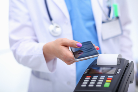 Doctor is holding payment terminal in hands. Paying for health care. Doctorの写真素材