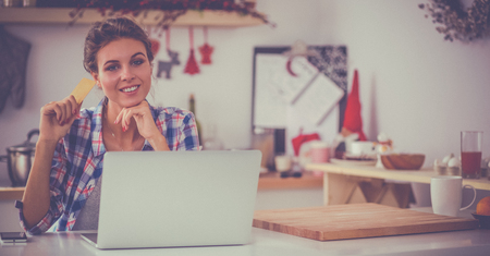 Smiling woman online shopping using computer and credit card in kitchenの写真素材