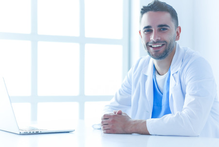 Portrait of a male doctor with laptop sitting at desk in medical officeの写真素材