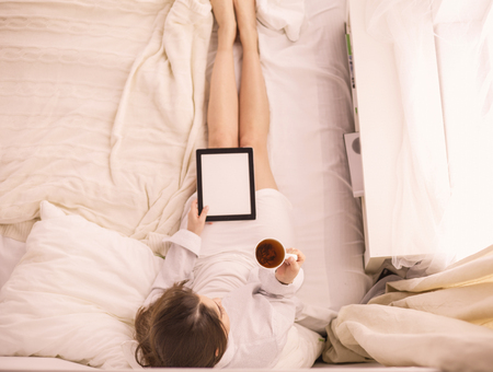 Portrait of woman using tablet and drinking coffee while sitting on the bed in the morningの写真素材