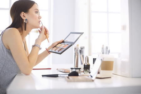A beautiful young woman sitting at a makeup table and doing her makeupの写真素材