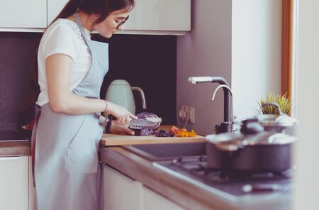 Young woman cutting vegetables in kitchen standing near deskの写真素材