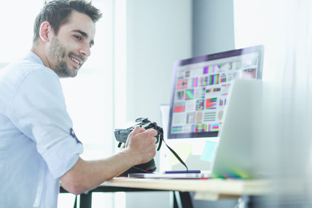 Portrait of young designer sitting at graphic studio in front of laptop and computer while working...