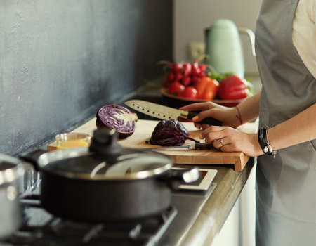 Young woman cutting vegetables in kitchen standing near deskの写真素材