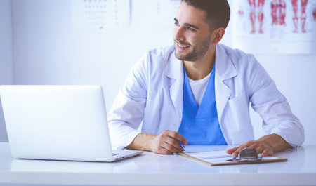 Portrait of a male doctor with laptop sitting at desk in medical officeの写真素材
