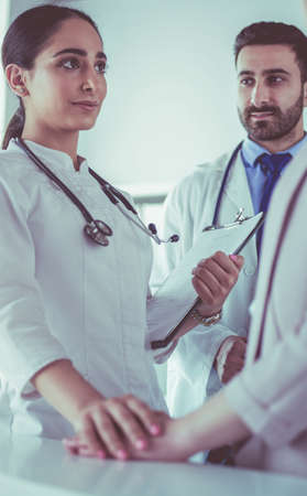 Handsome doctor is talking with young female patient and making notes while sitting in his officeの写真素材