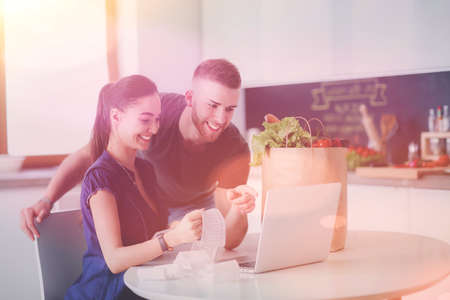 Young man cutting vegetables and woman standing with laptop in the kitchenの写真素材