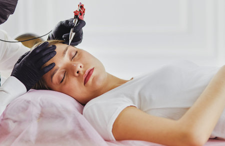 Young woman undergoing procedure of eyebrow permanent makeup in beauty salon