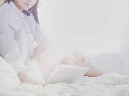 Portrait of woman using tablet and drinking coffee while sitting on the bed in the morningの写真素材