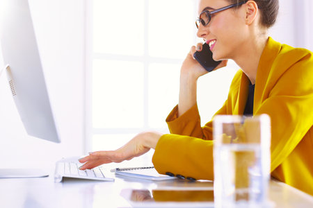 Businesswoman concentrating on work, using computer and cellphone in officeの写真素材