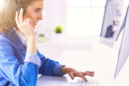 Happy charming young woman sitting and working with laptop using headset in officeの写真素材