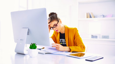 Attractive woman sitting at desk in office, working with laptop computer, holding documentの写真素材