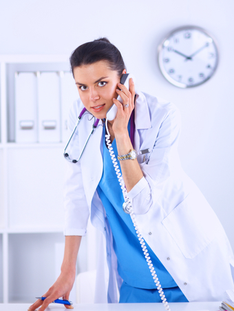 Portrait of young woman doctor in white coat at computer using phoneの写真素材