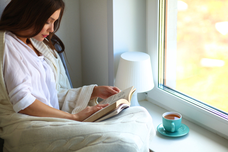Young woman at home sitting near window relaxing in her living room reading book and drinking coffee or tea .の写真素材