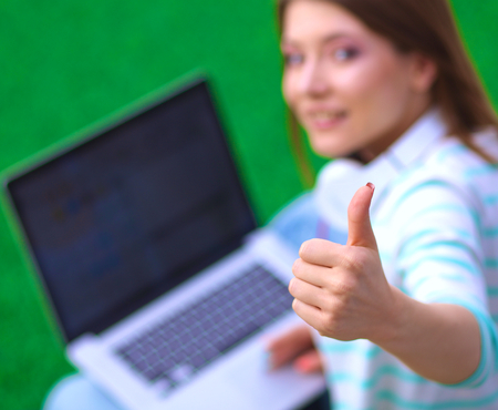 Young woman with laptop sitting on green grass and showing ok.の写真素材